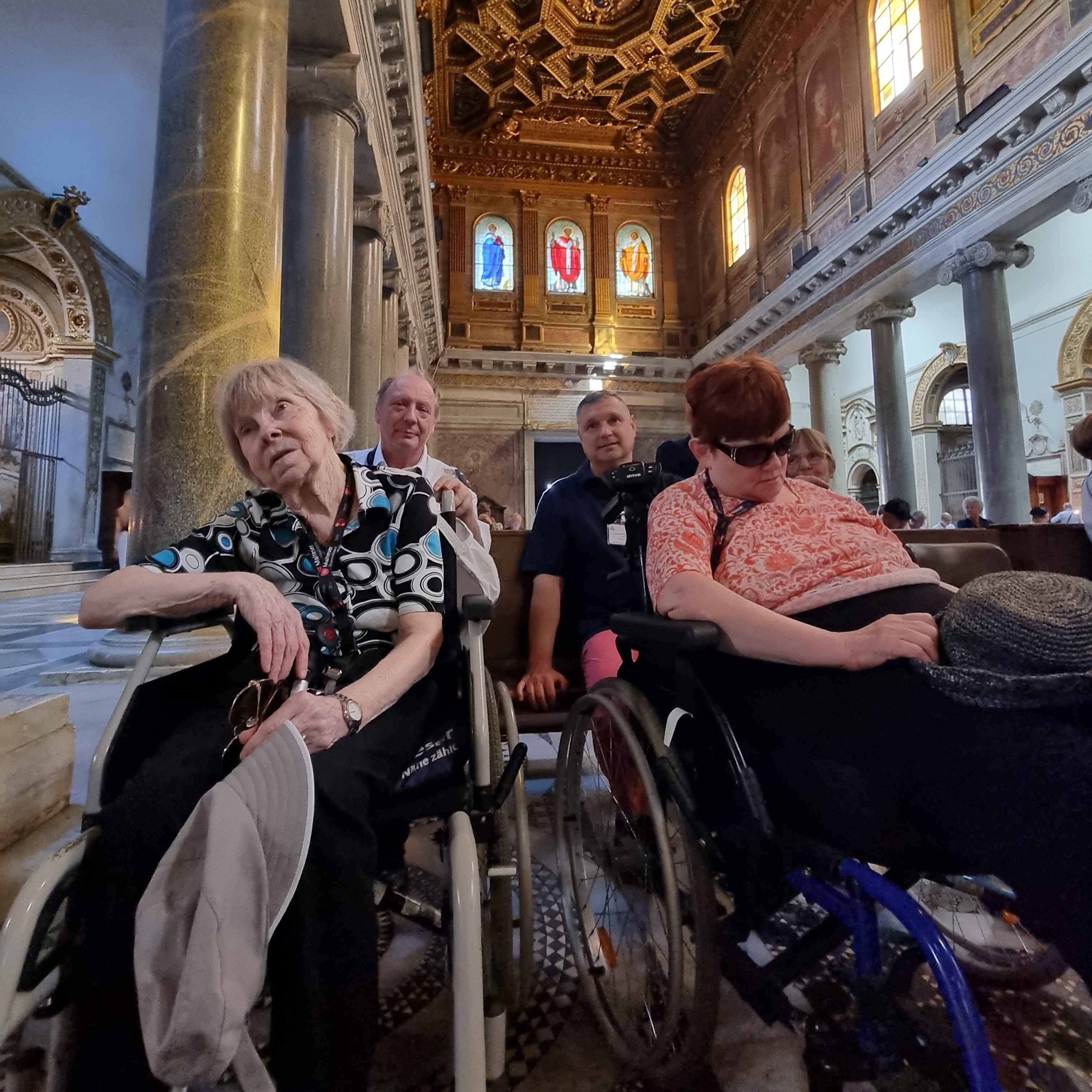 Vier Personen in Rollstühlen in einer Kirche mit hohen Säulen und bunten Glasfenstern im Hintergrund.