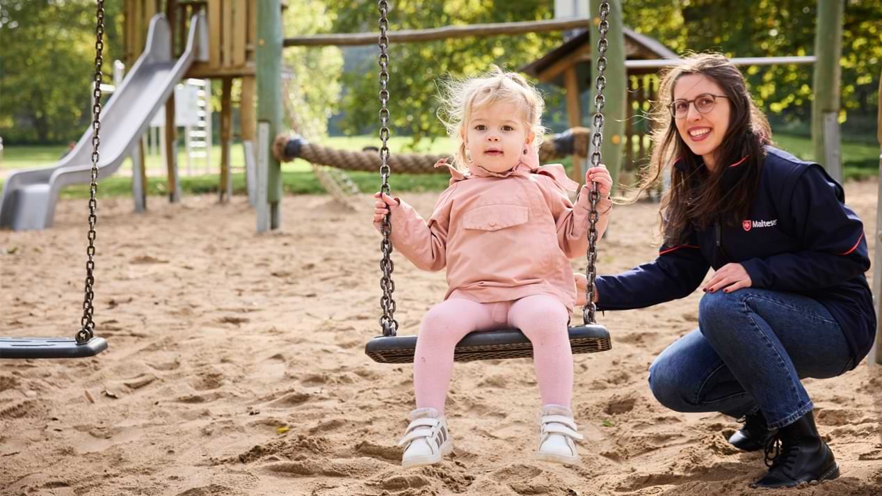 Ein Kleinkind in rosa Kleidung sitzt auf einer Schaukel auf einem Spielplatz. Neben ihm kniet eine erwachsene Frau mit Brille und dunkler Jacke und hält die Schaukel fest. Im Hintergrund sind Sandboden, Spielgeräte aus Holz, eine Rutsche und grüne Bäume zu sehen.
