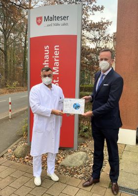 Arzt und Mann im Anzug stehen vor einem Malteser Schild und halten gemeinsam eine Urkunde.