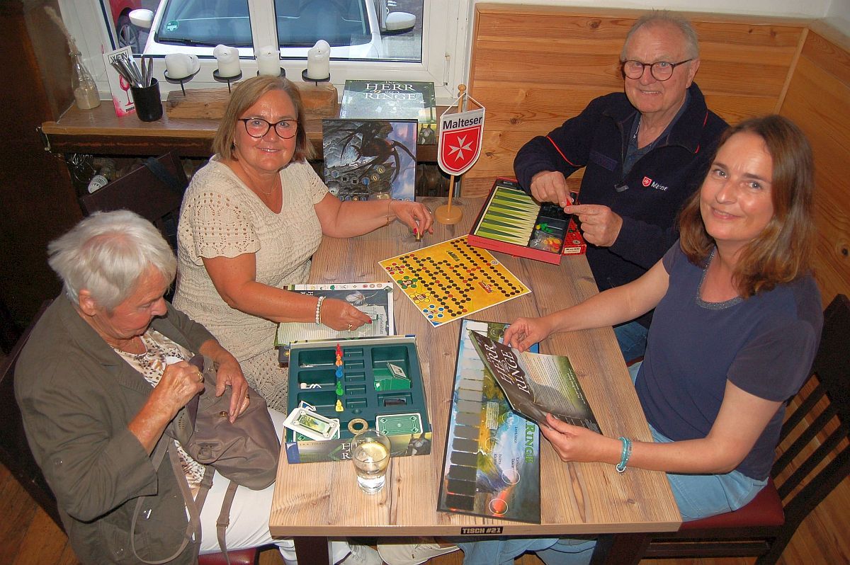 Auf dem Foto zu sehen sind vier Menschen, die gemeinsam am Tisch sitzen und ein Brettspiel vor sich haben. Sie lachen in die Kamera. Auf dem Tisch steht auch ein Wimpel der Malteser. 