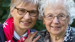 Frau mit Malteser Halstuch umarmt herzlich eine ältere Frau mit grauen Haaren, beide lächeln in die Kamera