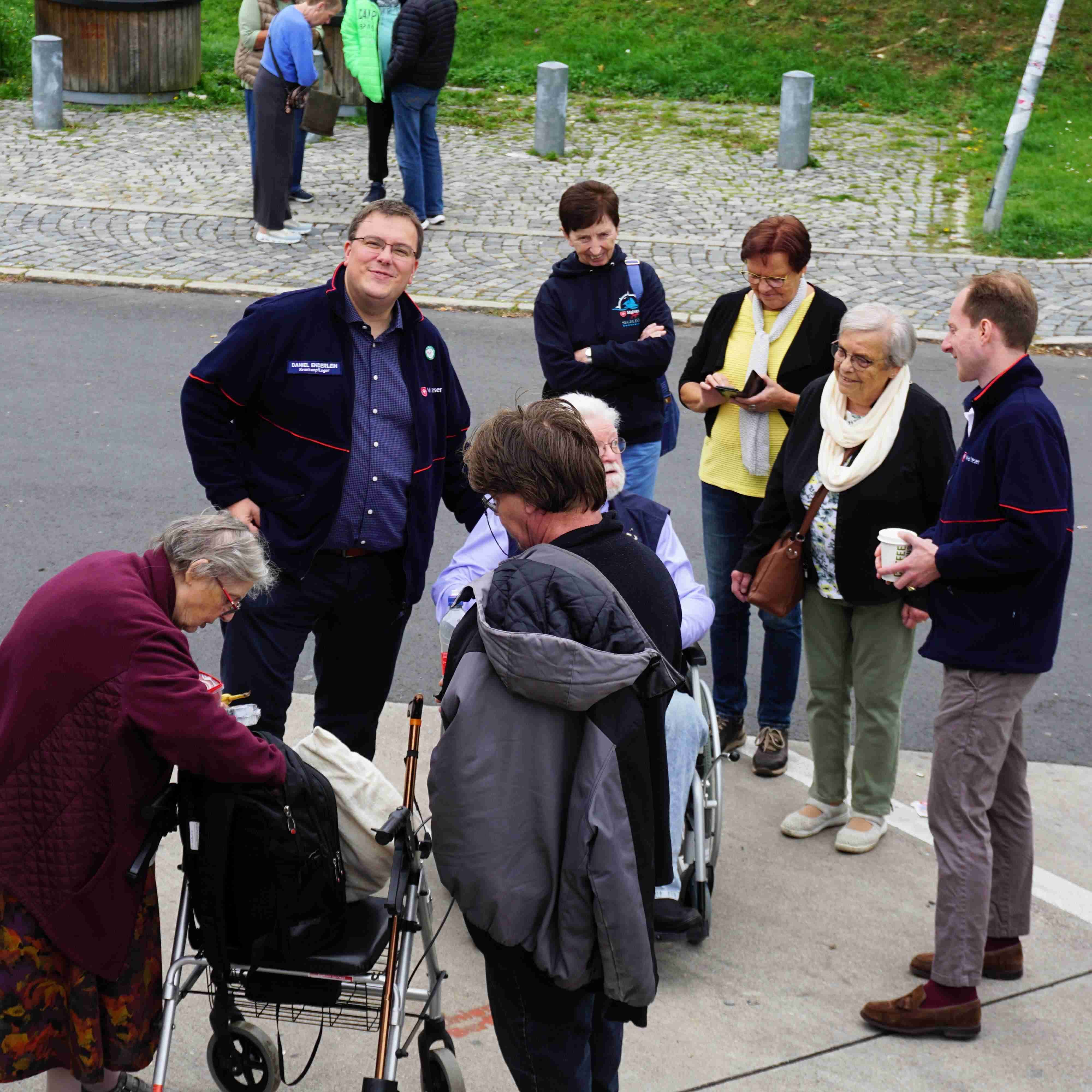 Gruppe von Menschen steht auf Gehweg und Straße, darunter eine Person im Rollstuhl und eine ältere Frau mit Rollator.