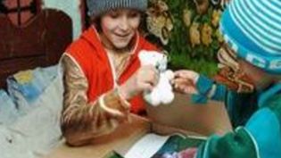 Warm angezogene Kinder packen ein Paket aus, das Kind mit der grauen Mütze hält einen weißen Teddybären in der Hand