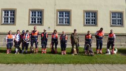 12 Männer und Frauen und einige Hunde stehen aufgereiht vor einem Kasernengebäude.