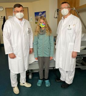 Zwei Ärzte und ein Kind mit Maske stehen in einem Patientenzimmer vor einem Klinikbett.