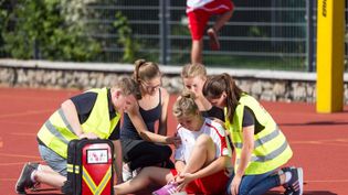 Schülerinnen und Schüler in gelben Warnwesten knien um ein am Bein verletztes Mädchen auf einem roten Sportplatz