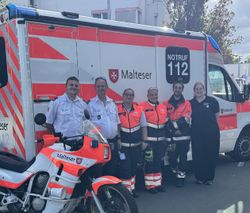 Sechs Malteser stehen in Dienstkleidung nebeneinander vor einem Rettungswagen, schräg davor steht ein Motorrad, ebenfalls im Malteser Design.