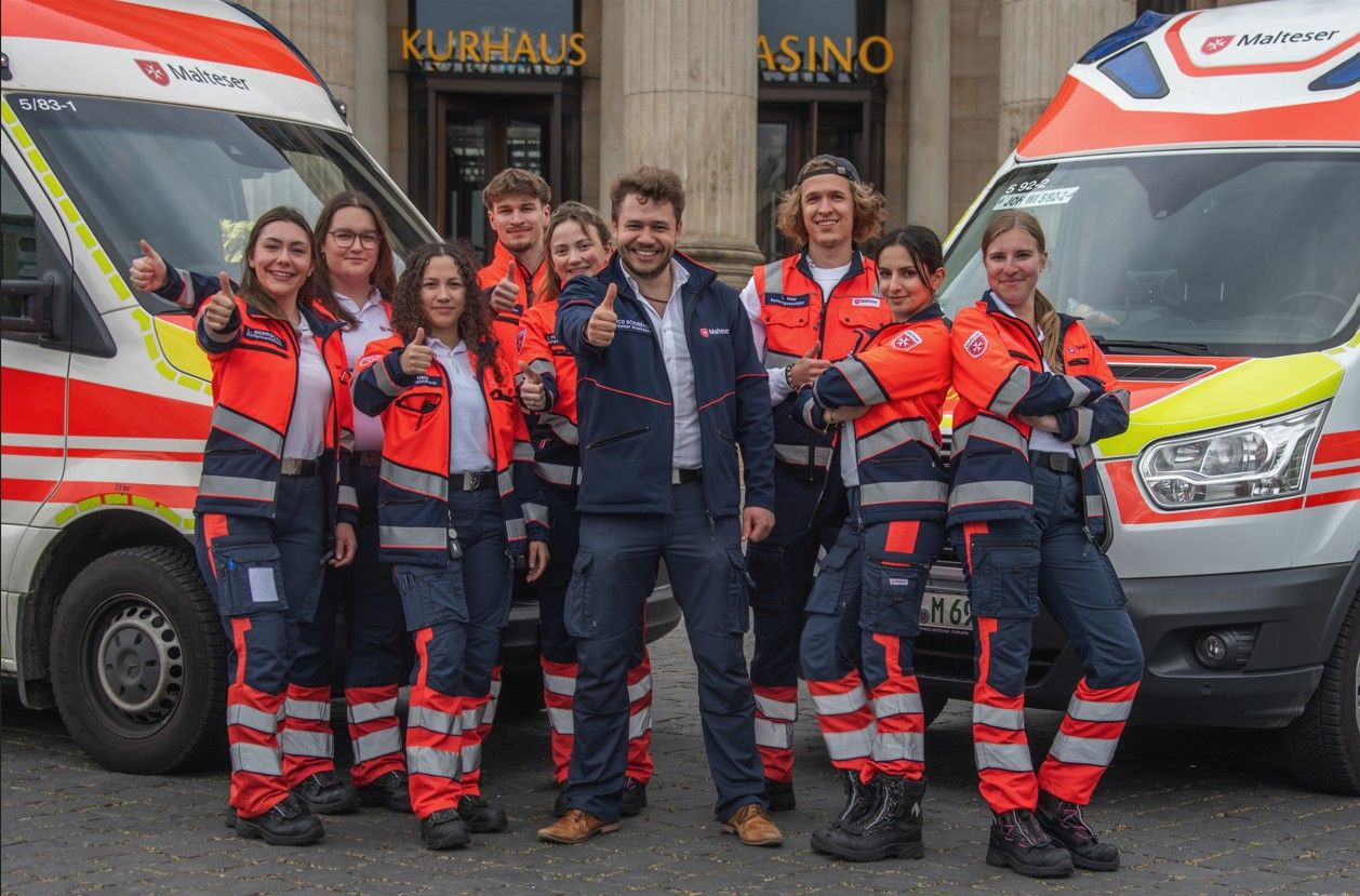 Ein Team aus Notfallsanitäterinnen und Rettungssanitätern der Malteser Wiesbaden steht vor dem Wiesbadener Kurhaus. Neben ihnen befinden sich ein Rettungswagen und ein Krankentransportwagen.