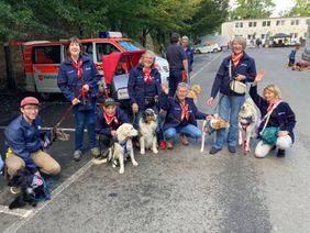 Auf dem Foto sieht man mehrere Personen des Besuchs- und Begleitungsdienstes mit Herz und Hund zusammen mit Ihren Hunden. Im Hintergrund ist ein Einsatzfahrzeug der Malteser sowie weitere Personen zu erkennen.