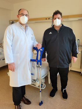 Arzt in weißem Kittel steht mit maskiertem Patienten im Patientenzimmer, beide halten gemeinsam Gehhilfen in der Hand.