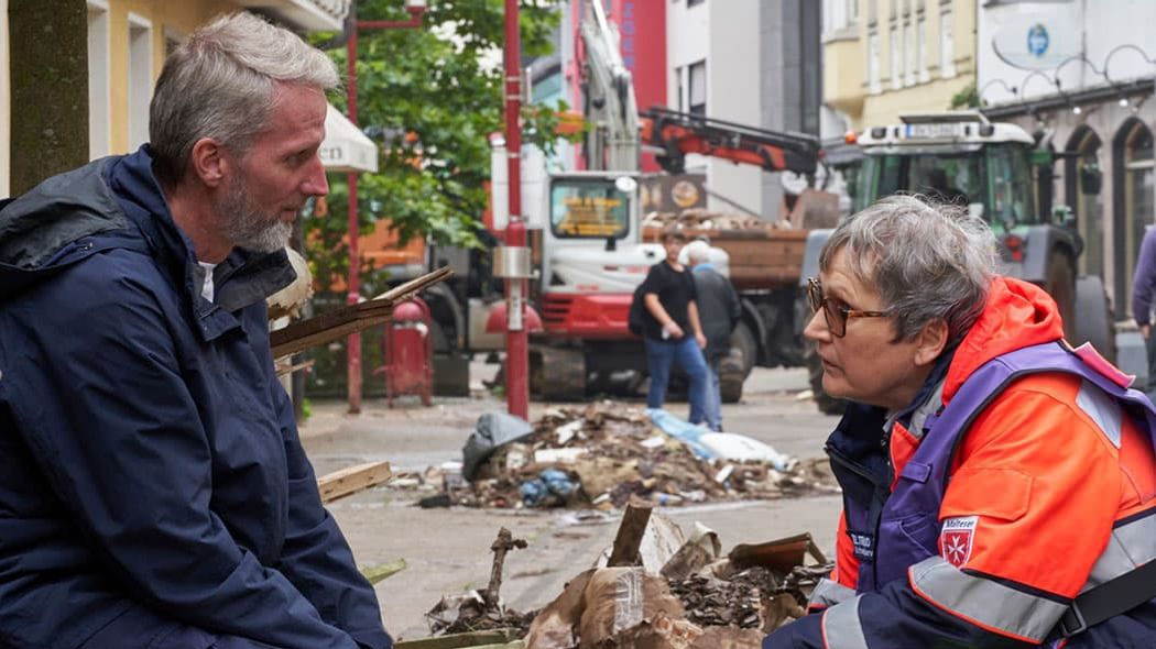 Malteser Mitarbeiterin in Einsatzkleidung im Gespräch mit einem Betroffenen der Flutkatastrophe.