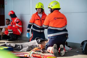 Zwei Malteser helfen einem Verletzten, der am Boden liegt.