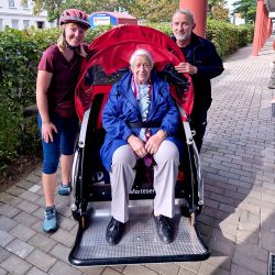 Eine Frau sitzt in einer Fahrradrikscha. Neben ihr stehen eine Frau und ein Mann.