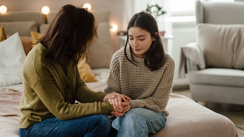Zwei Frauen sitzen auf einer Bettkante und halten sich an den Händen.