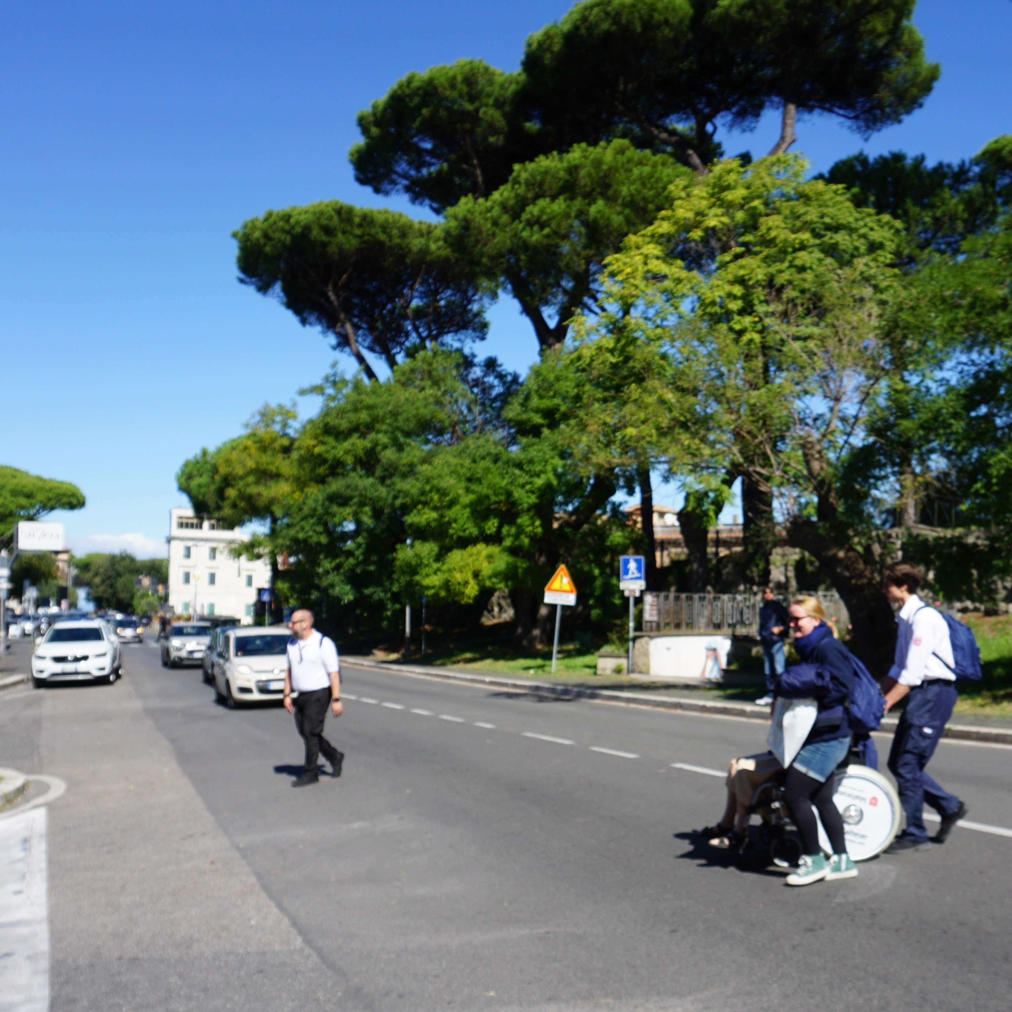 Straße in Castel Gandolfo mit mehreren Autos, Fußgänger und Bäumen mit breiten Kronen unter blauem Himmel.