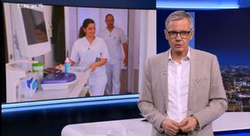 Nachrichtensprecher vor Bildschirm, auf dem Pflegende in Klinikflur gezeigt werden.