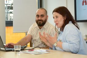 Die zwei Projektmitarbeitenden Maen Al Darwish (links) und Buschra Al-Ani (rechts) sitzen an einem Tisch. Frau Al-Ani spricht und gestikuliert dabei, Herr Al Darwish sieht sie dabei an und lächelt.