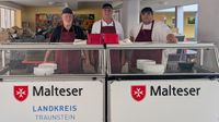 Drei Malteser Helfer stehen hinter einer Theke zur Essensausgabe.
