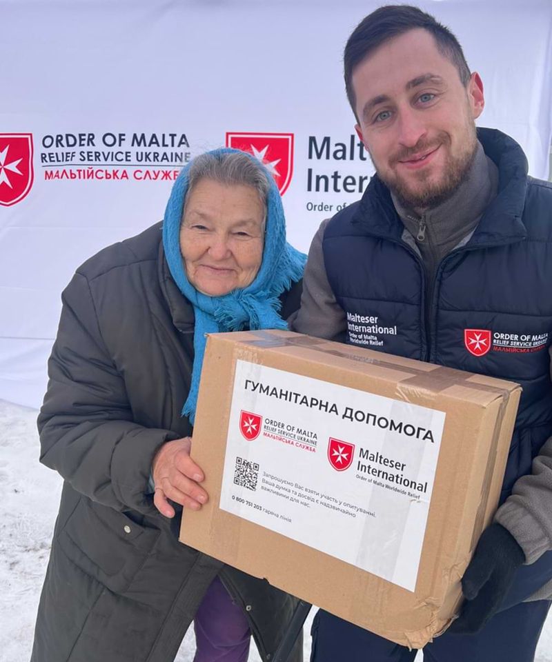Ein lächelnder Mann mit einer Daunenweste übergibt ein Hilfspaket an eine lächelnde alte Frau in Winterkleidung.