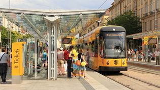 Straßenbahn an moderner Haltestelle in Dresden mit wartenden Fahrgästen und überdachtem Bahnsteig.
