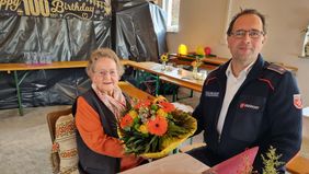 Die Jubilarin sitzt mit einem Blumenstrauß in der Hand an einem Tisch, daneben sitzt Malteser Kreisgeschäftsführer Roland Bucher