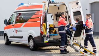 Malteser in Einsatzkleidung helfen einem gehbehindertem Mann in in ein Fahrzeug.