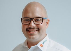 Ein 30 Jahre alter Mann lächelt in die Kamera. Er trägt ein Hemd mit Malteser-Beschriftung.