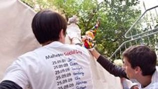 Zwei Jungen malen etwas auf ein Plakat. Sie tragen weiße T-Shirts mit der Aufschrift Social Day.