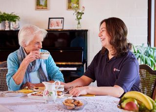 Eine junge Frau im Malteser Shirt trinkt mit einer alten Frau Kaffee
