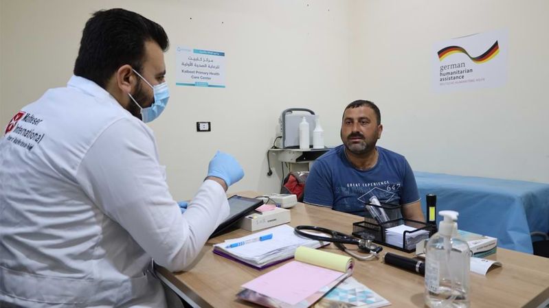 Ein Arzt mit Mund-Nasen-Schutz bei einem Patientengespräch an einem Tisch.