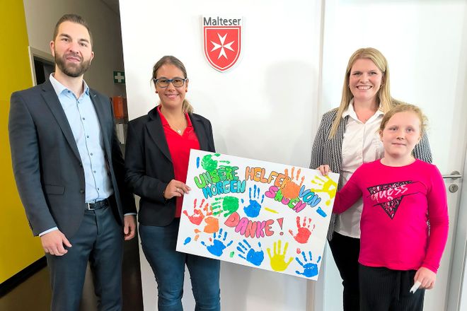 Vier Personen stehen in einem Innenraum und halten gemeinsam ein bunt bemaltes Bild mit Handabdrücken und Zeichnungen. Im Hintergrund ist ein Wappen an der Wand zu sehen. Die Gruppe präsentiert das Bild für die Kamera.