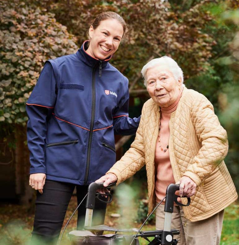 Eine junge Frau in Malteser Jacke und eine ältere Dame mit Rollator stehen im Grünen und lächeln.