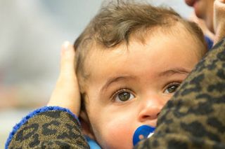 Kleines Kind mit Schnuller im Mund blick mit großen traurigen Augen über die Schulter seiner Mutter, die es behutsam auf dem Arm trägt. 