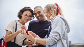 Drei junge Malteser Mitarbeitende, jeglichen Aussehens und Geschlechts, schauen lächelnd auf ein Handy, dass sie gemeinsam mittig vor sich halten.