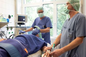 Anästhesiepersonal setzt einem Patienten eine Beatmungsmaske auf, im Hintergrund ein Beatmungsgerät und Anschlüsse.