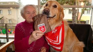 Eine ältere Frau sitzt mit einem Golden Retriever mit rotem Halstuch auf einem Sessel.
