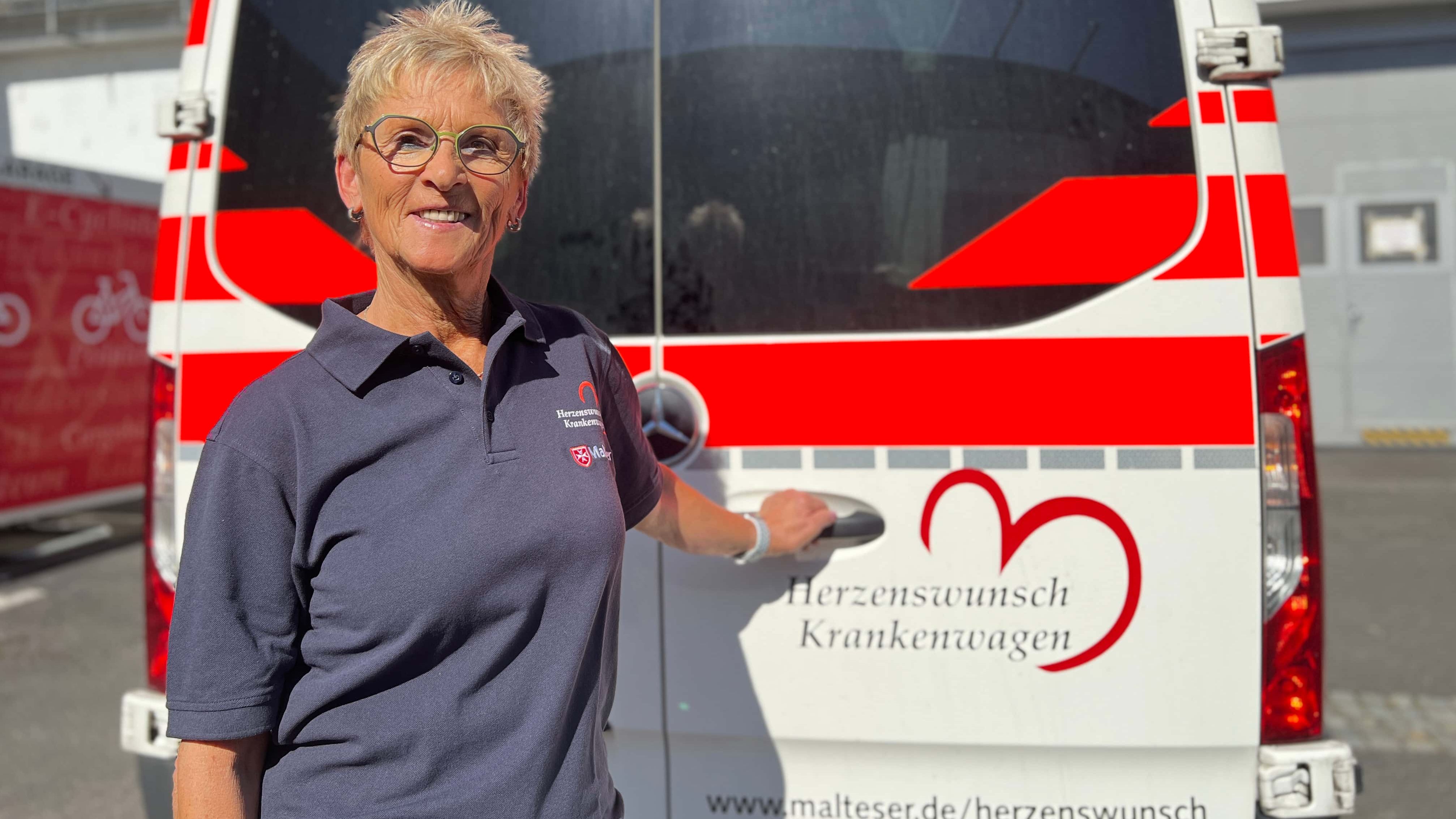 Eine Frau steht vor einem Malteser Herzenswunsch Krankenwagen und hält die Hecktür fest.