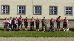 12 Männer und Frauen und einige Hunde stehen aufgereiht vor einem Kasernengebäude.