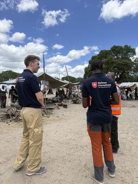 Drei Männer im Gespräch, dahinter Menschen unter einfachen zelten. Foto: Malteser International