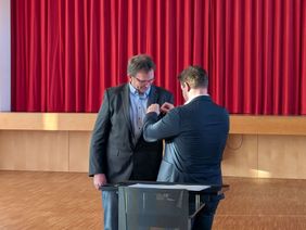 Ein Mann steckt einem anderen eine Ehrennadel an. 