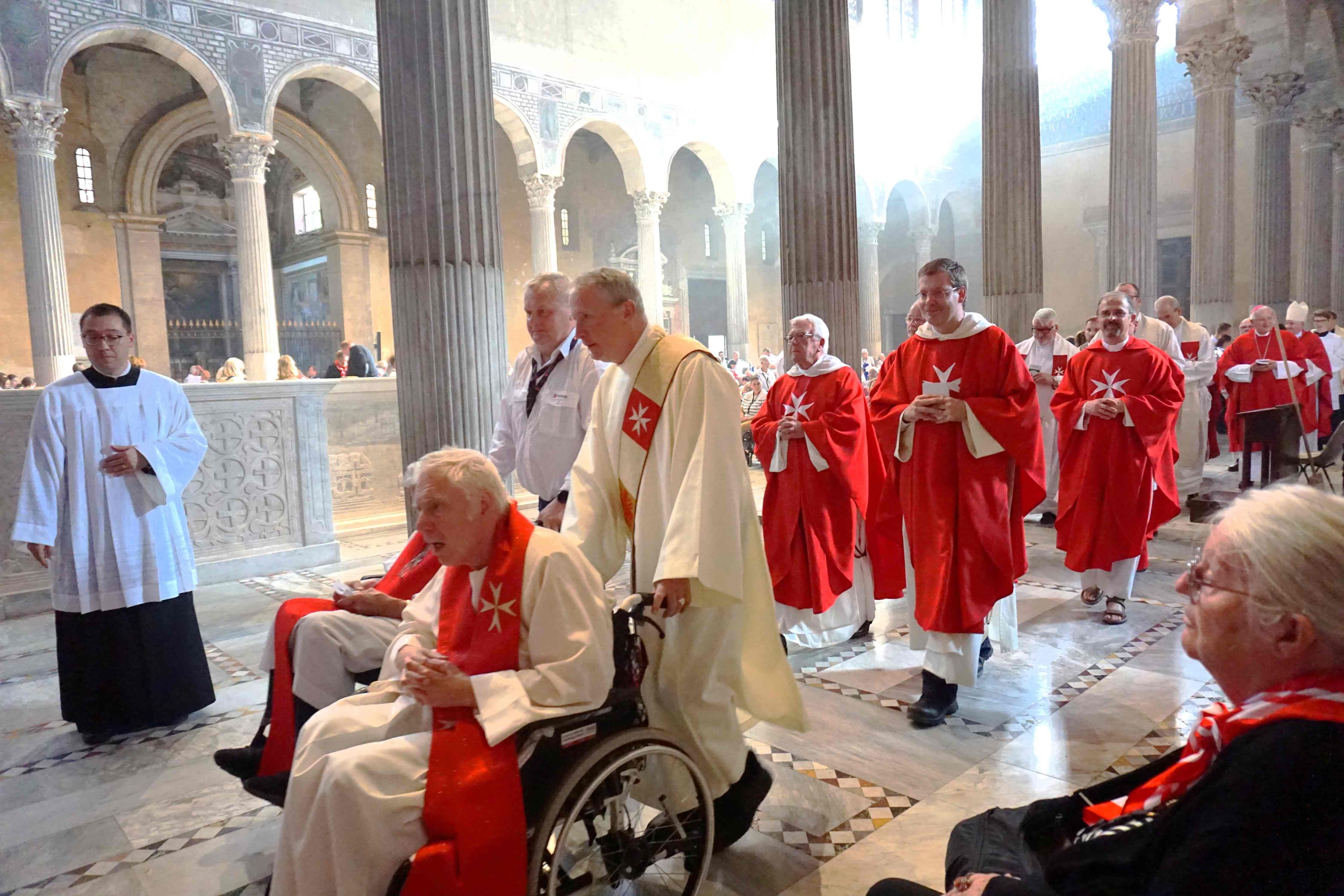 In der Kirche laufen mehrere Menschen in liturgischen Gewändern mit Malteser Kreuz. Am Anfang der Reihe werden zwei Menschen in Rollstühlen geschoben.