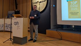 Mann hält einen Vortrag bei einer Veranstaltung, im Hintergrund ist eine Präsentation zu sehen.