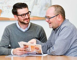 Zwei Männer sitzen am Tisch und schauen eine Broschüre durch und unterhalten sich dabei