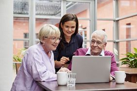 Eine ältere Frau und ein älterer Mann sitzen am Laptop. Zwischen beiden steht eine junge Frau der Malteser, die ihnen etwas erklärt. (Foto: Malteser) 