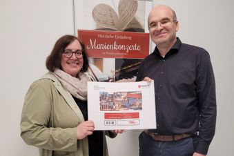 Frau und Mann halten gemeinsam einen Spendenscheck, im Hintergrund ein Plakat zu einer Konzertreihe.