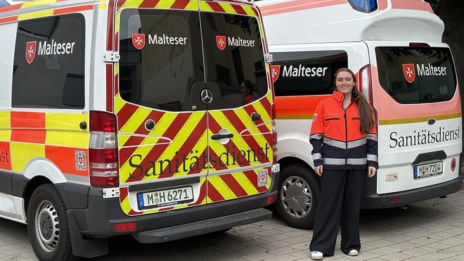 Eine junge Frau in Malteser Dienstkleidung steht zwischen zwei Einsatzfahrzeugen.