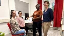 In einem Büro bekommen zwei Frauen, auf Stühlen sitzend, Gutscheine für Drogerieartikel und Lebensmittel von zwei stehenden Malteser Mitarbeiterinnen überreicht.