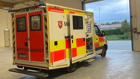 Ein Malteser Rettungswagen steht in der Fahrzeughalle mit geöffnetem Tor und Blick nach draußen.