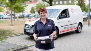 Mitarbeiterin des Malteser Menueservice trägt eine Warmhaltebox und lächelt in die Kamera.