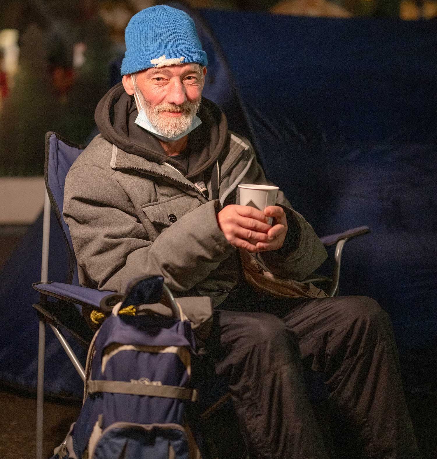 Ein älterer Mann mit Winterjacke sitzt in einem Campingstuhl mit einer Tasse in den Händen